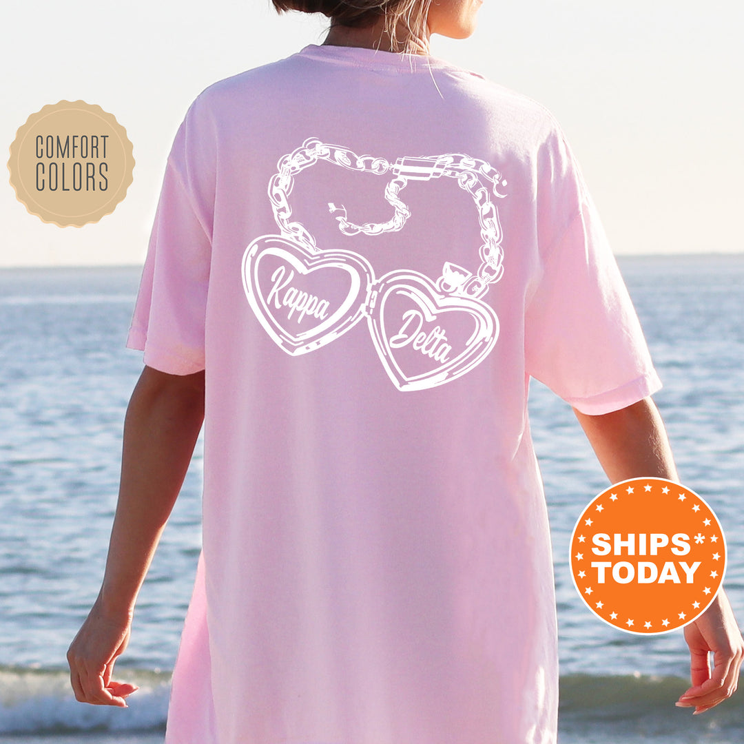 A person wearing a pink t-shirt with a graphic design of two heart-shaped charms on the back, standing near the ocean.