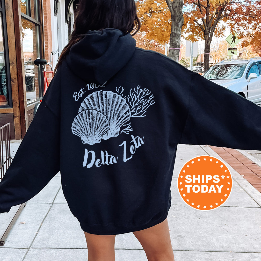 A person wearing a black hoodie with a white graphic of a shell and the text "Delta Zeta" printed on it, standing on a sidewalk.