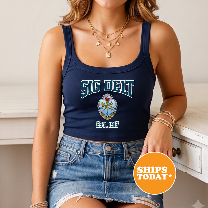 A woman wearing a navy blue crop top with the text "SIG DELT" and a navy blue skirt, along with multiple necklaces. She is sitting on a white surface, and there is a circular orange sign with the text "SHIPS TODAY" in the image.
