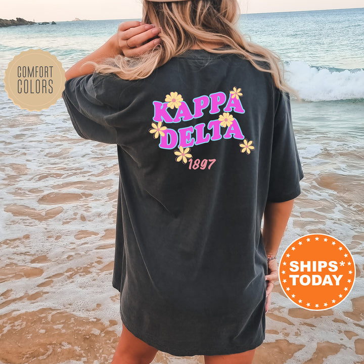 A woman wearing a black t-shirt with the text "KAPPADELTA" printed on it, standing on a beach with waves in the background.