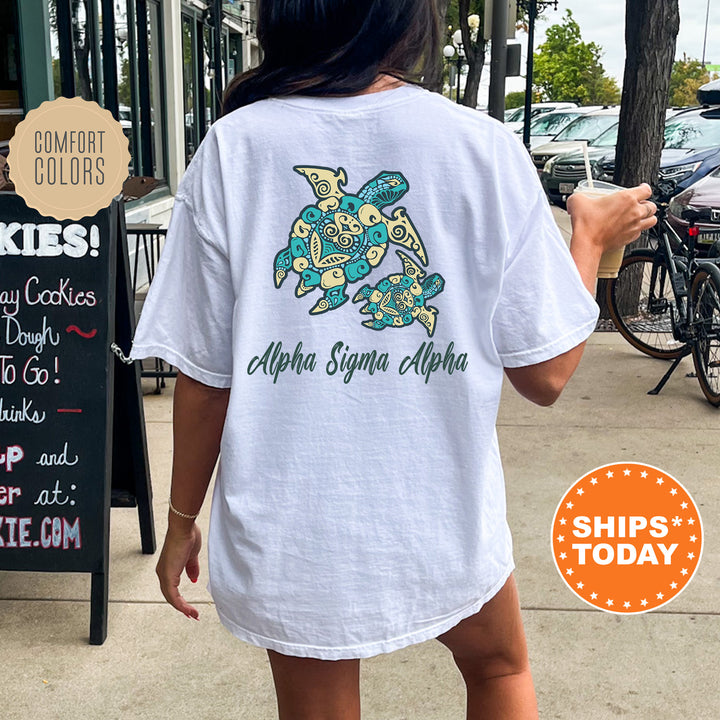 A person wearing a white t-shirt with a graphic of a turtle and text that says "Alpha Sigma Alpha" is standing on a sidewalk.