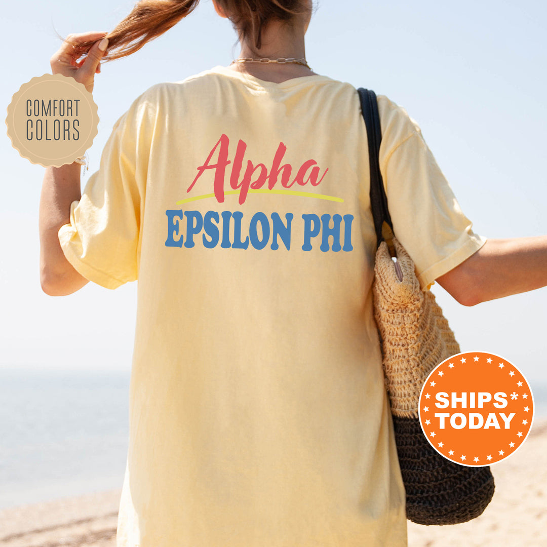 A person wearing a yellow t-shirt with the text "Alpha Epsilon Phi" printed on it, standing on a beach with a handbag.