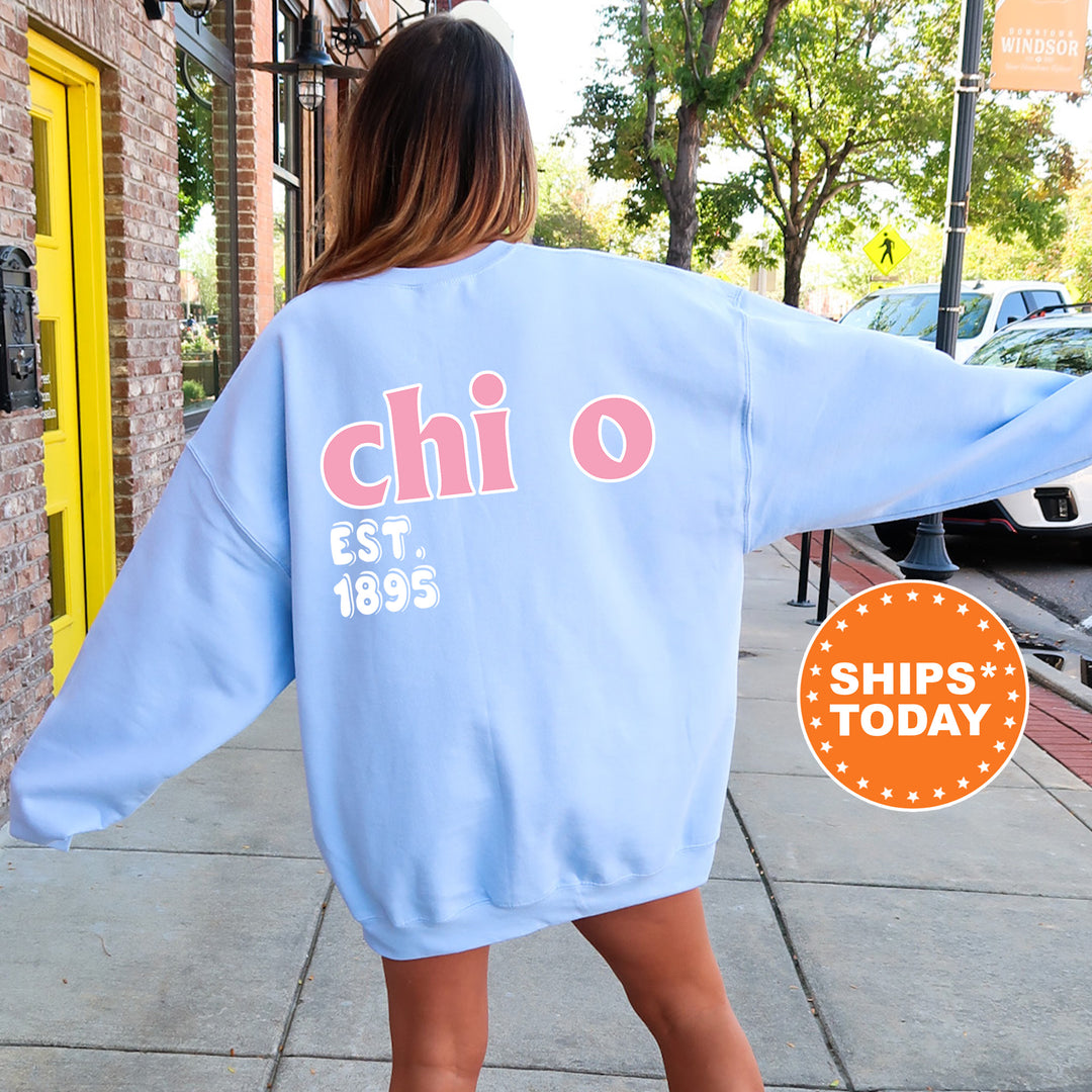 A woman wearing a light blue sweatshirt with the text "chi" and "EST. 1895" printed on it, standing on a sidewalk.