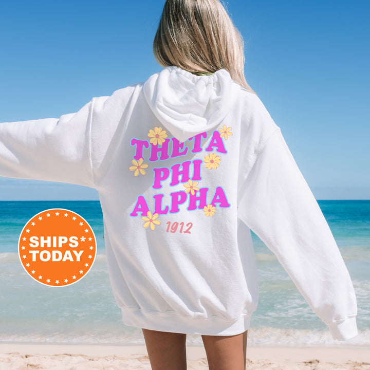 A woman wearing a white hoodie with the text "THETA PHI ALPHA 1912" printed on it, standing on a beach with the ocean in the background.
