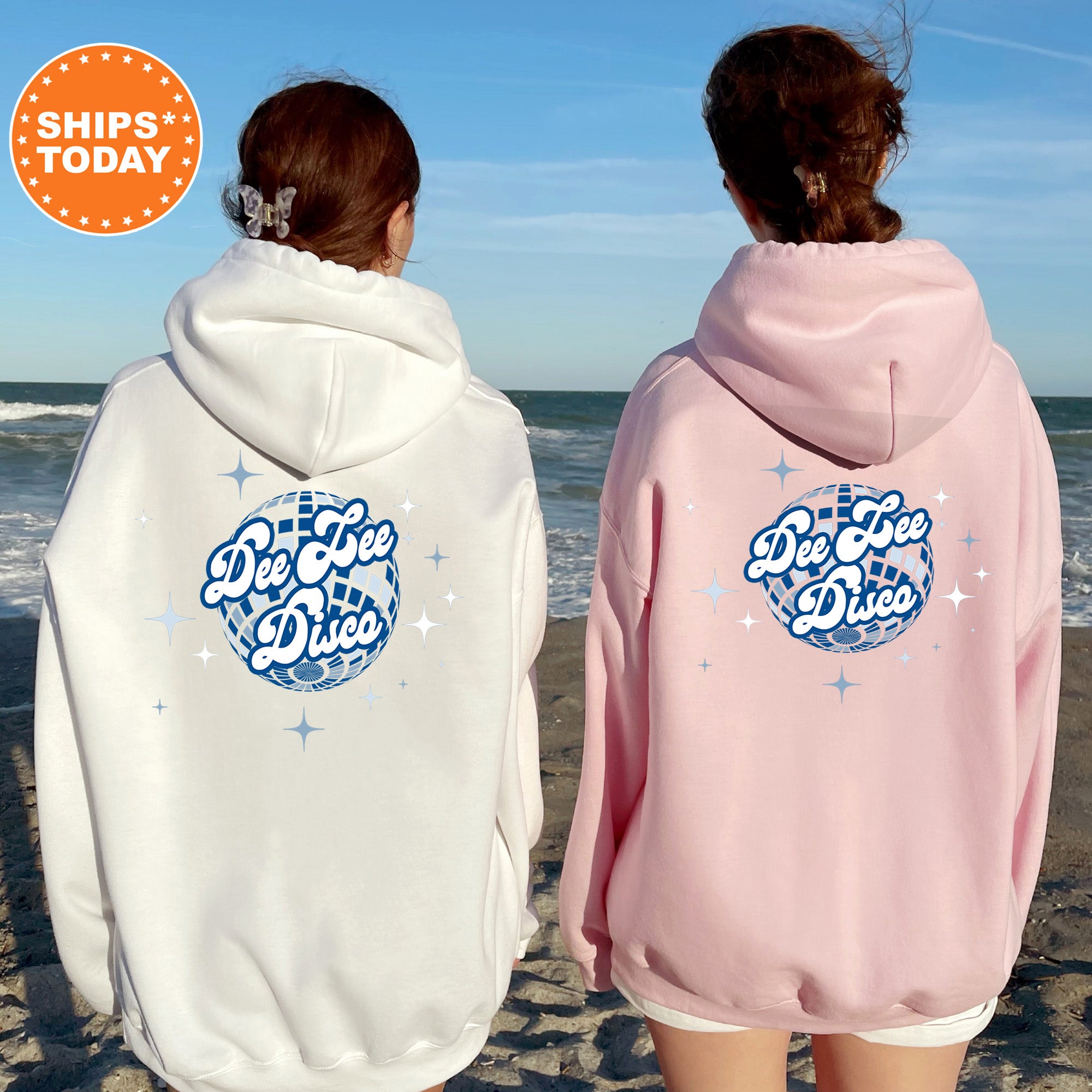 Two women standing on a beach, wearing hooded sweatshirts with the text 