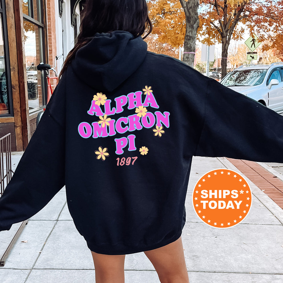 A person wearing a black hoodie with pink and yellow text and flowers on the back, standing on a sidewalk with a car and a building in the background.