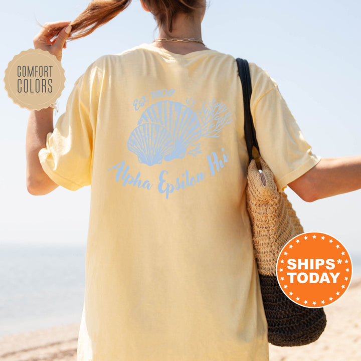 A person wearing a yellow t-shirt with a blue design and text, standing on a beach with a handbag and a sign advertising "SHIPS TODAY".
