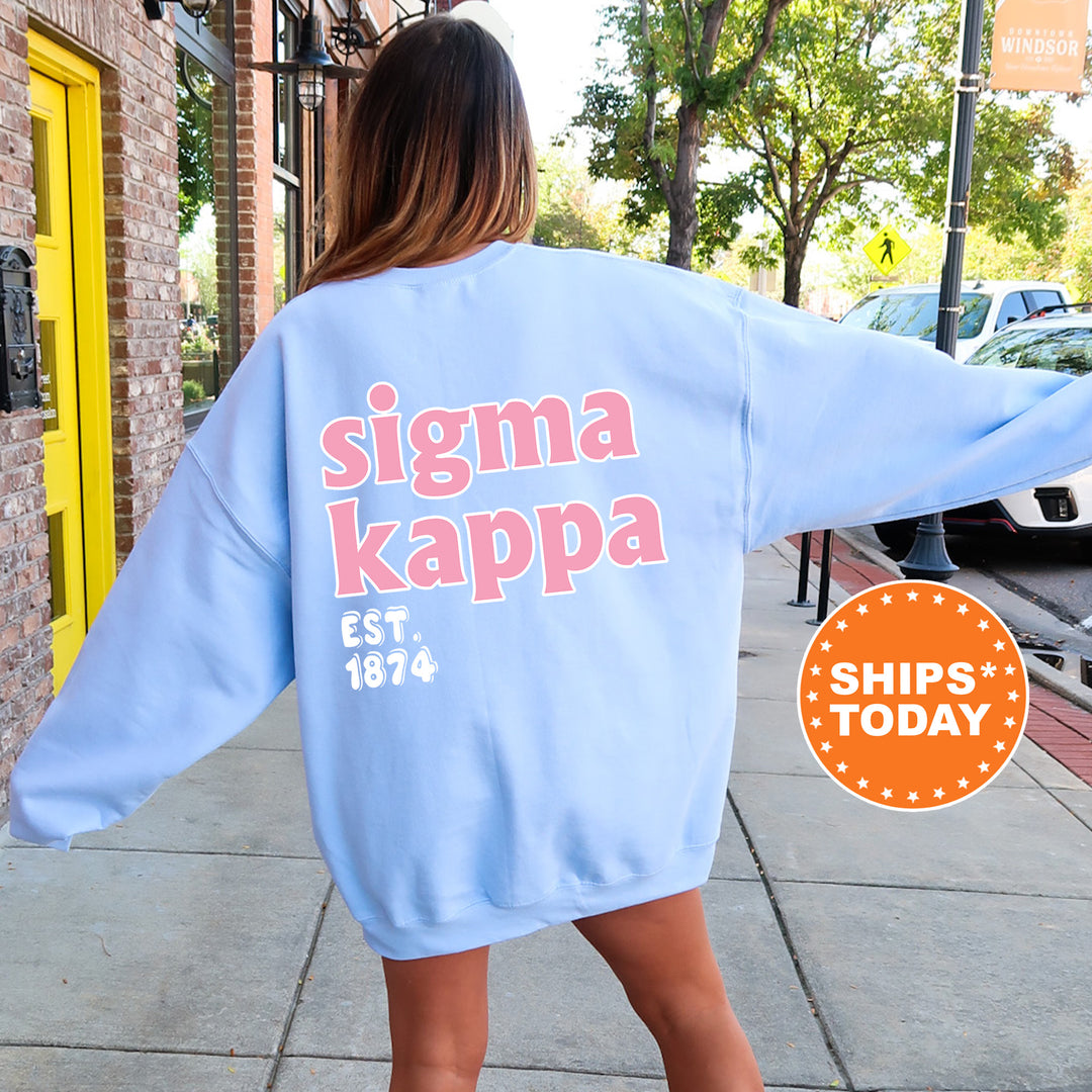 A woman wearing a light blue oversized sweatshirt with the text " Sigma Kappa EST. 1874" printed on it, standing on a sidewalk.