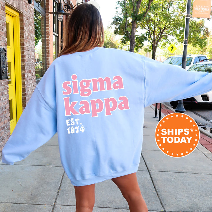A woman wearing a light blue oversized sweatshirt with the text " Sigma Kappa EST. 1874" printed on it, standing on a sidewalk.