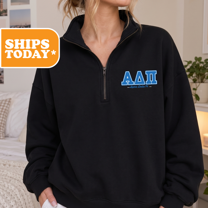A person wearing a black sweatshirt with the letters 'AA' and 'ΑΓΑΙΚΑΙ' printed on it, standing in a room with a bed and a plant in the background.