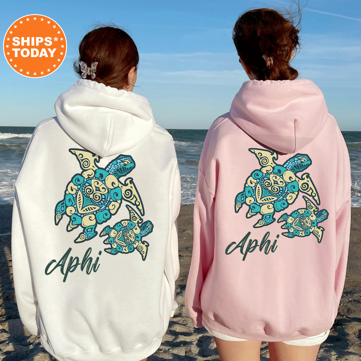 Two women wearing hooded sweatshirts with a turtle design on the back, standing on a beach with the ocean in the background.