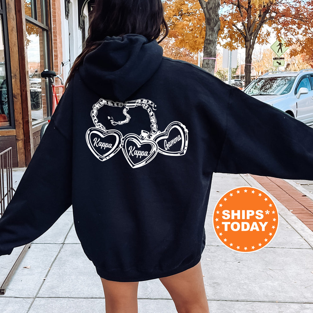 A person wearing a black hoodie with a graphic design on the back, standing on a sidewalk with a car and trees in the background.