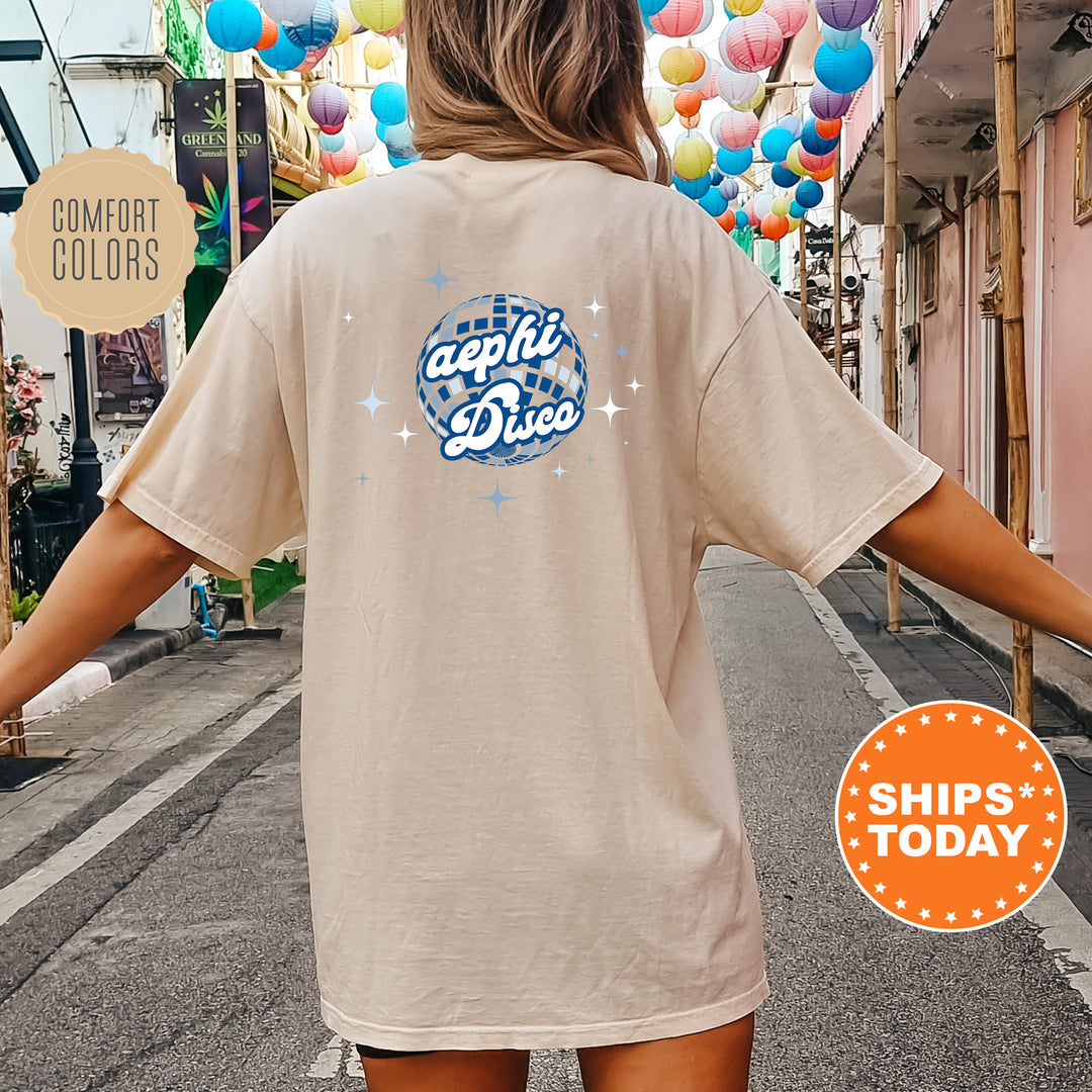 A person wearing a beige t-shirt with a graphic design on the back, standing on a street with colorful lanterns hanging overhead.