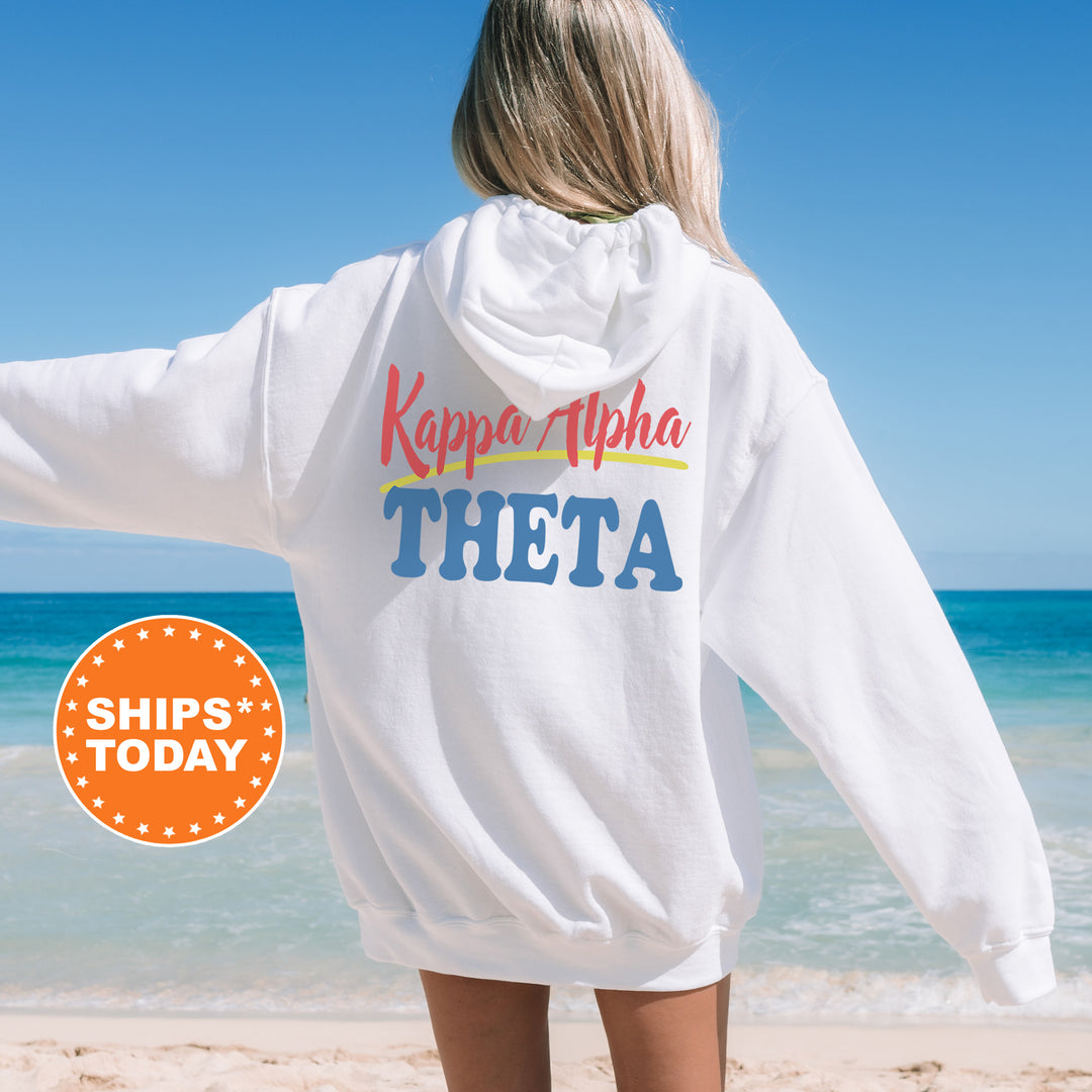 A woman wearing a white hoodie with the text " Kappa Alpha Theta" printed on it, standing on a beach with the ocean in the background.