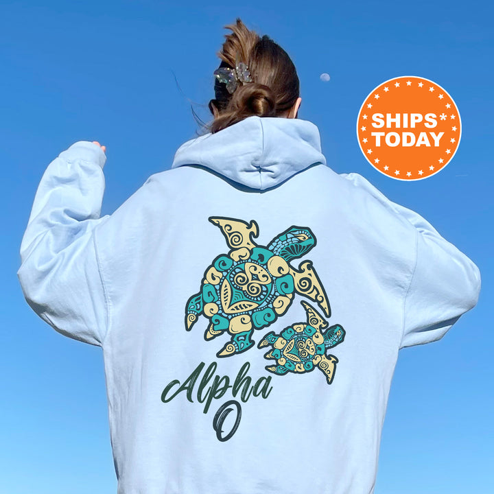 A person wearing a light blue hoodie with a colorful turtle design and the word "Alpha" printed on it, standing against a clear blue sky.