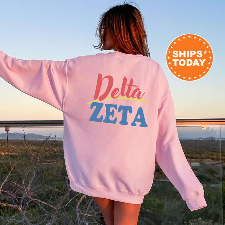 A woman wearing a pink sweatshirt with the text "Delta Zeta" printed on it, standing in front of a scenic outdoor landscape with mountains in the background.