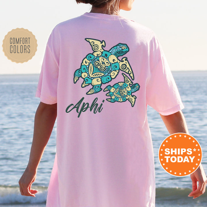 A person wearing a pink t-shirt with a graphic of a turtle and the word "Aphi" printed on it, standing near the ocean.