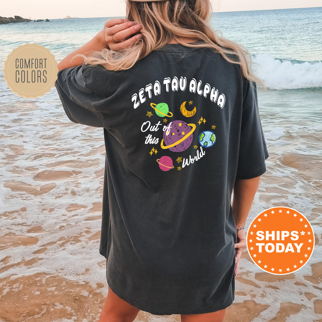 A woman wearing a black t-shirt with a graphic design of planets and stars on the back, standing on a beach with the ocean in the background.
