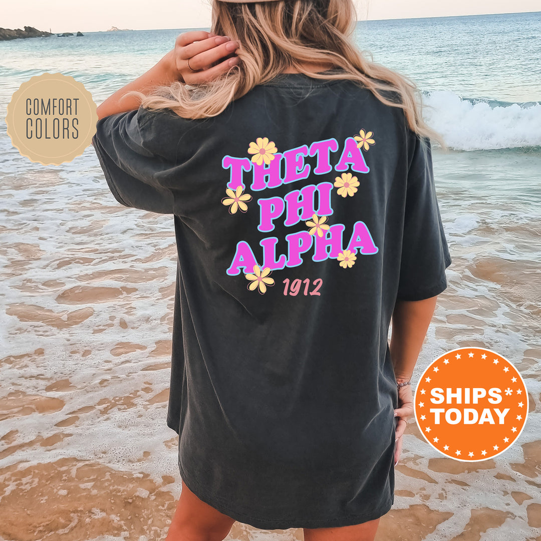 A woman wearing a black t-shirt with the text "THETA PHI ALPHA 1912" printed on it, standing on a beach with the ocean in the background.