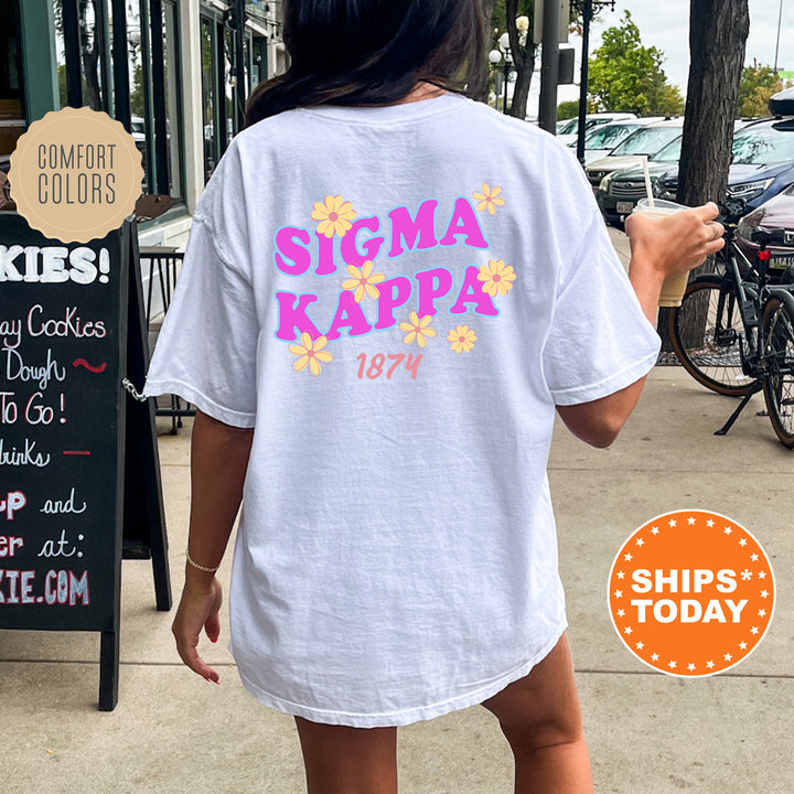 A person wearing a white t-shirt with the text "SIGMA KAPPAS" and "1874" printed on it, standing on a sidewalk next to a chalkboard sign.