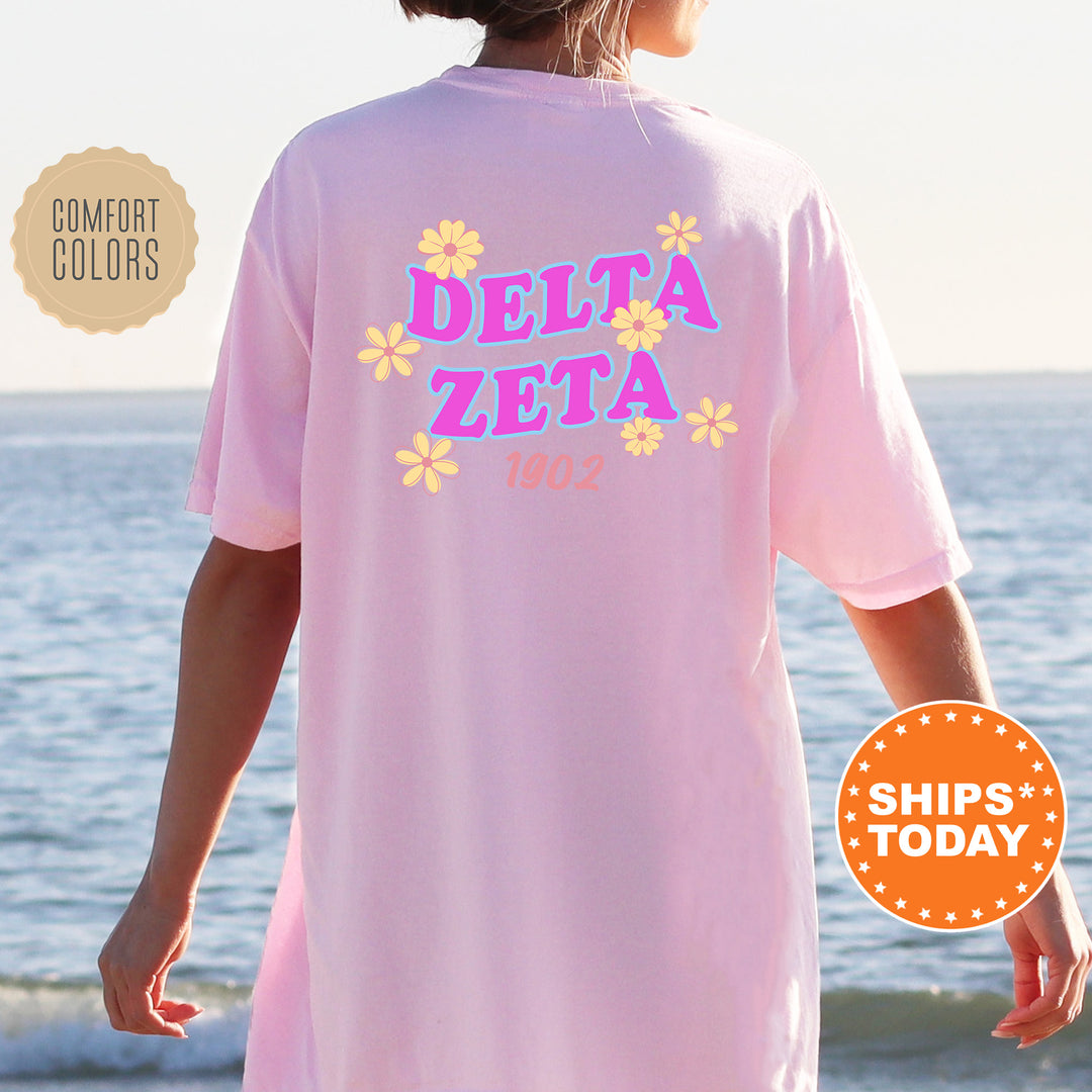 A person wearing a pink t-shirt with the text "DELTA ZETA" and "1902" printed on it, standing near the ocean.