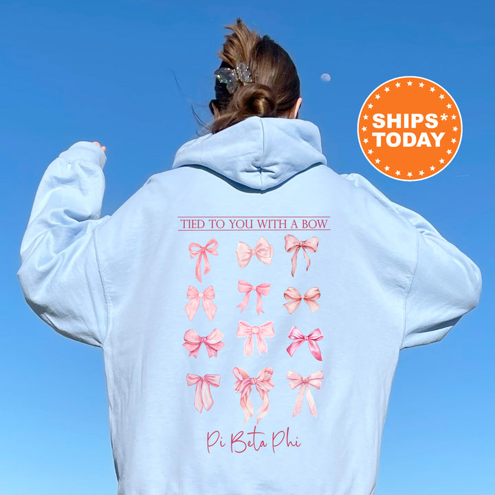 A person wearing a light blue hoodie with a graphic of pink bows on the back, standing against a clear blue sky.
