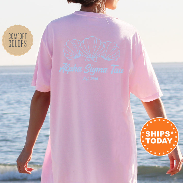 A person wearing a pink t-shirt with the text "Alpha Sigma Tau" and "Est. 1998" printed on it, standing on a beach with the ocean in the background.