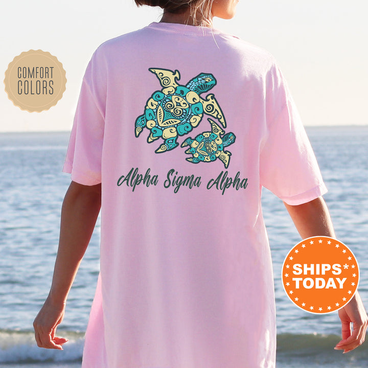 A person wearing a pink t-shirt with a graphic of a turtle and the text "Alpha Sigma Alpha" printed on it, standing near the ocean.