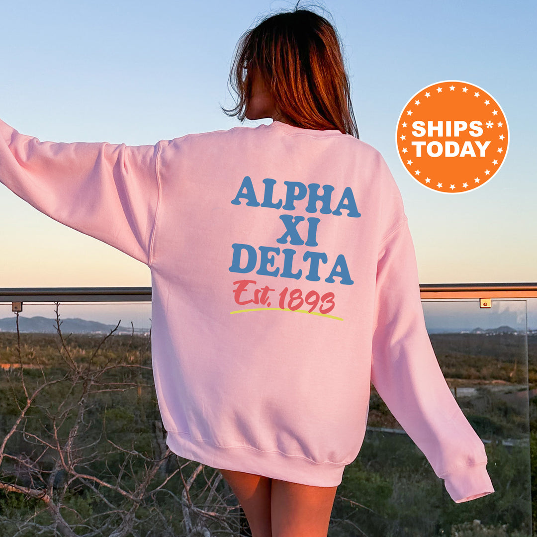 A woman wearing a pink sweatshirt with the text "ALPA XI DELTA Est.1893" printed on it, standing in front of a scenic outdoor landscape.