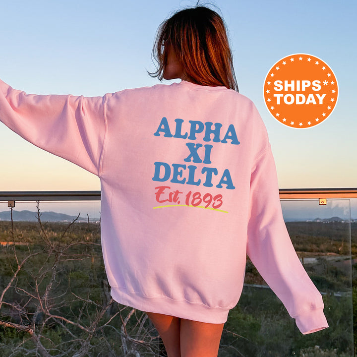 A woman wearing a pink sweatshirt with the text "ALPA XI DELTA Est.1893" printed on it, standing in front of a scenic outdoor landscape.