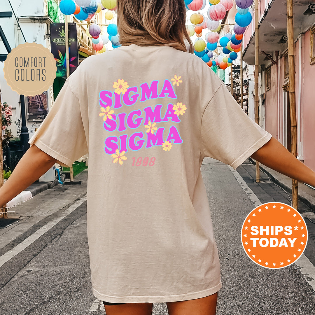 A person wearing a beige t-shirt with the text "SIGMA SIGMA SIGMA" printed on it, standing in front of a street lined with colorful lanterns.