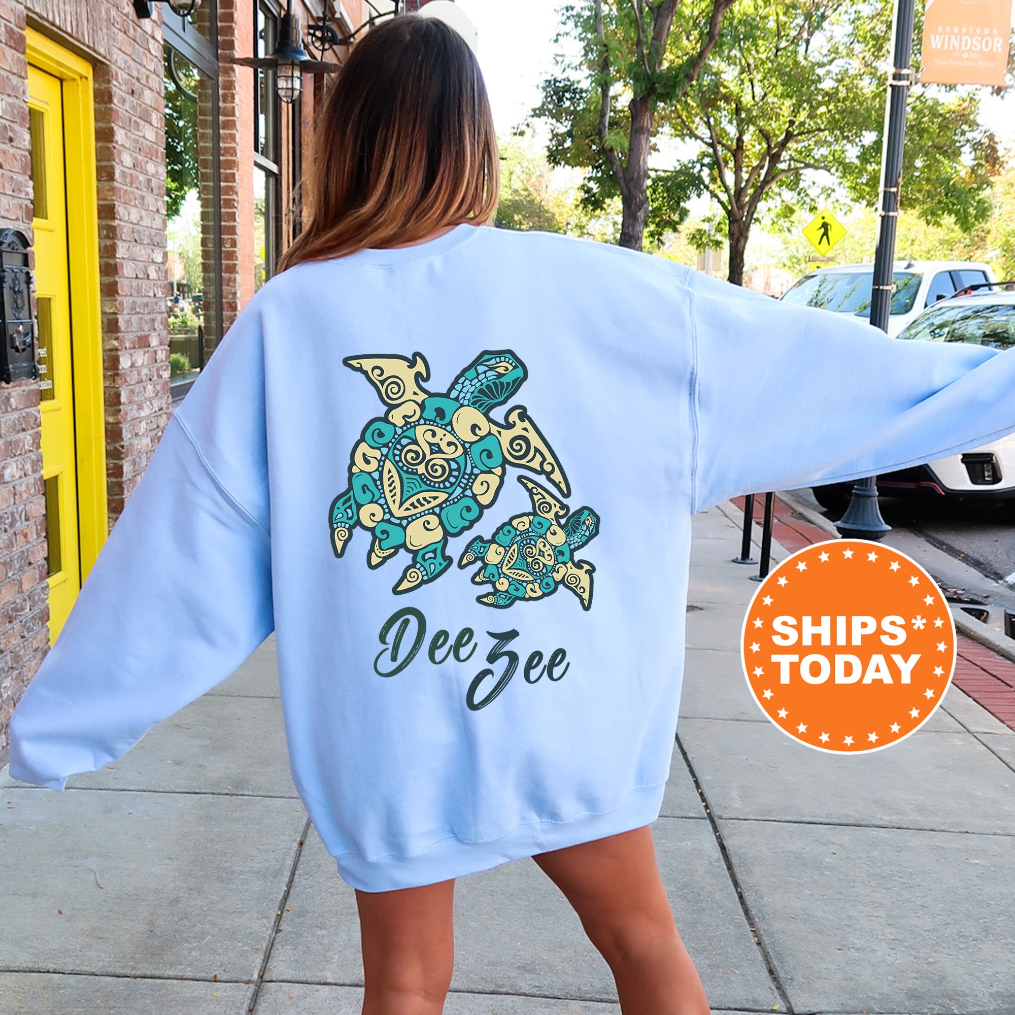 A woman wearing a light blue sweatshirt with a design of a turtle and the name 'Dee' printed on it, standing on a sidewalk.