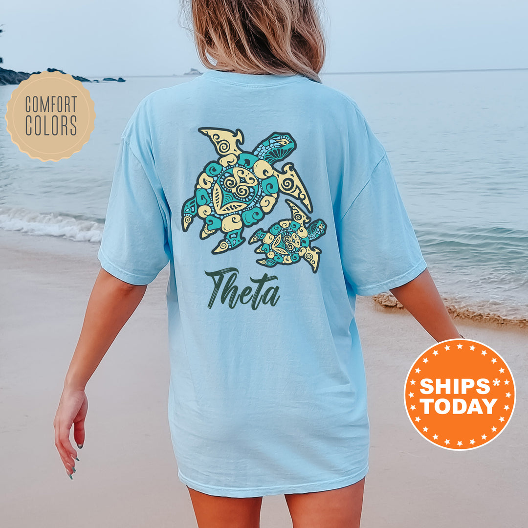 A woman wearing a light blue t-shirt with a design of two turtles on the back, standing on a beach with the ocean in the background.