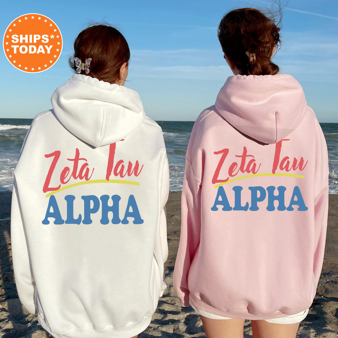 Two women wearing matching white and pink hoodies with the text "Zeta Jau ALPHA" printed on them, standing on a beach with the ocean in the background.