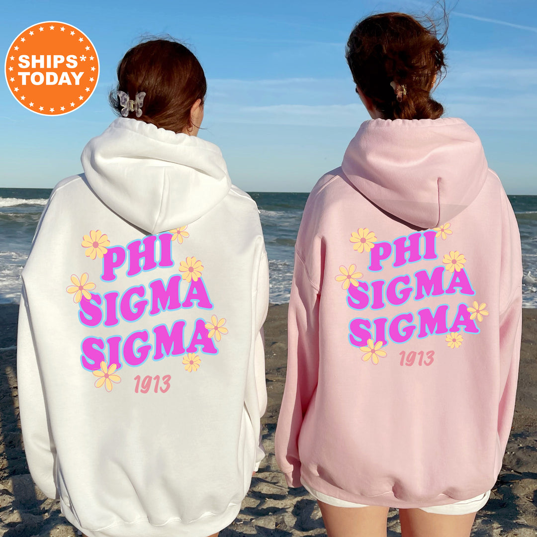 Two women wearing hooded sweatshirts with the text "PHI SIGMA SIGMA" printed on them, standing on a beach with the ocean in the background.