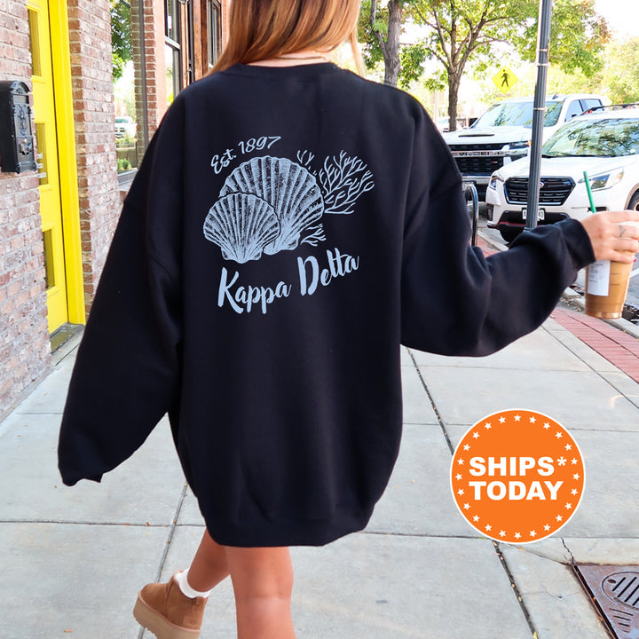 A person wearing a black sweatshirt with a graphic design of seashells and the text " Kappa Delta" printed on it, walking on a sidewalk.