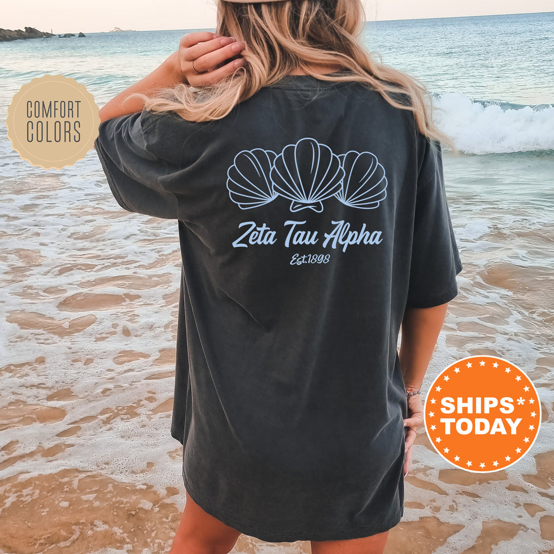 A woman wearing a black t-shirt with a graphic design of three shells on the back, standing on a beach with the ocean in the background.