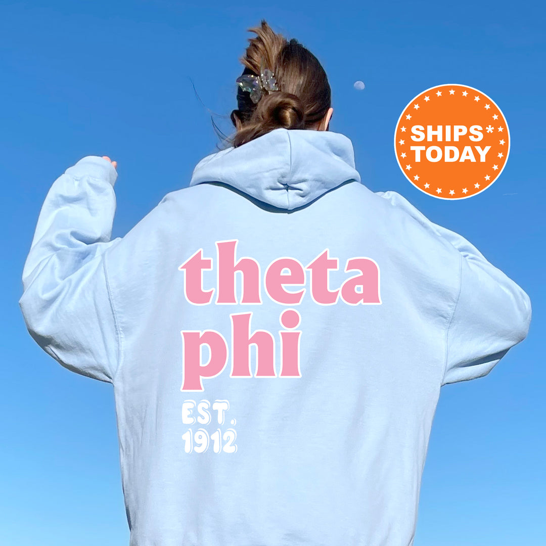 A person wearing a light blue hoodie with the text "theta phi" printed on it, standing against a clear blue sky.