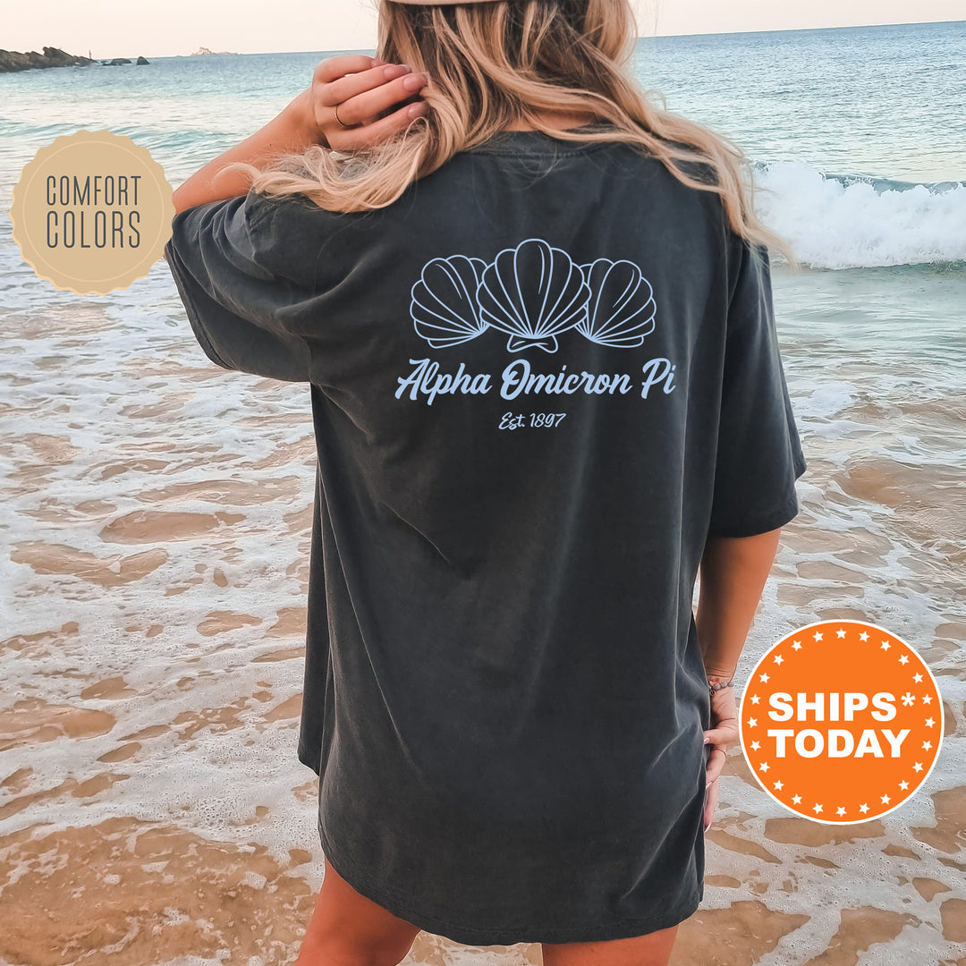 A woman wearing a black t-shirt with a graphic design of seashells and the text "Alpha Omega Pi" on the back, standing on a beach with the ocean in the background.