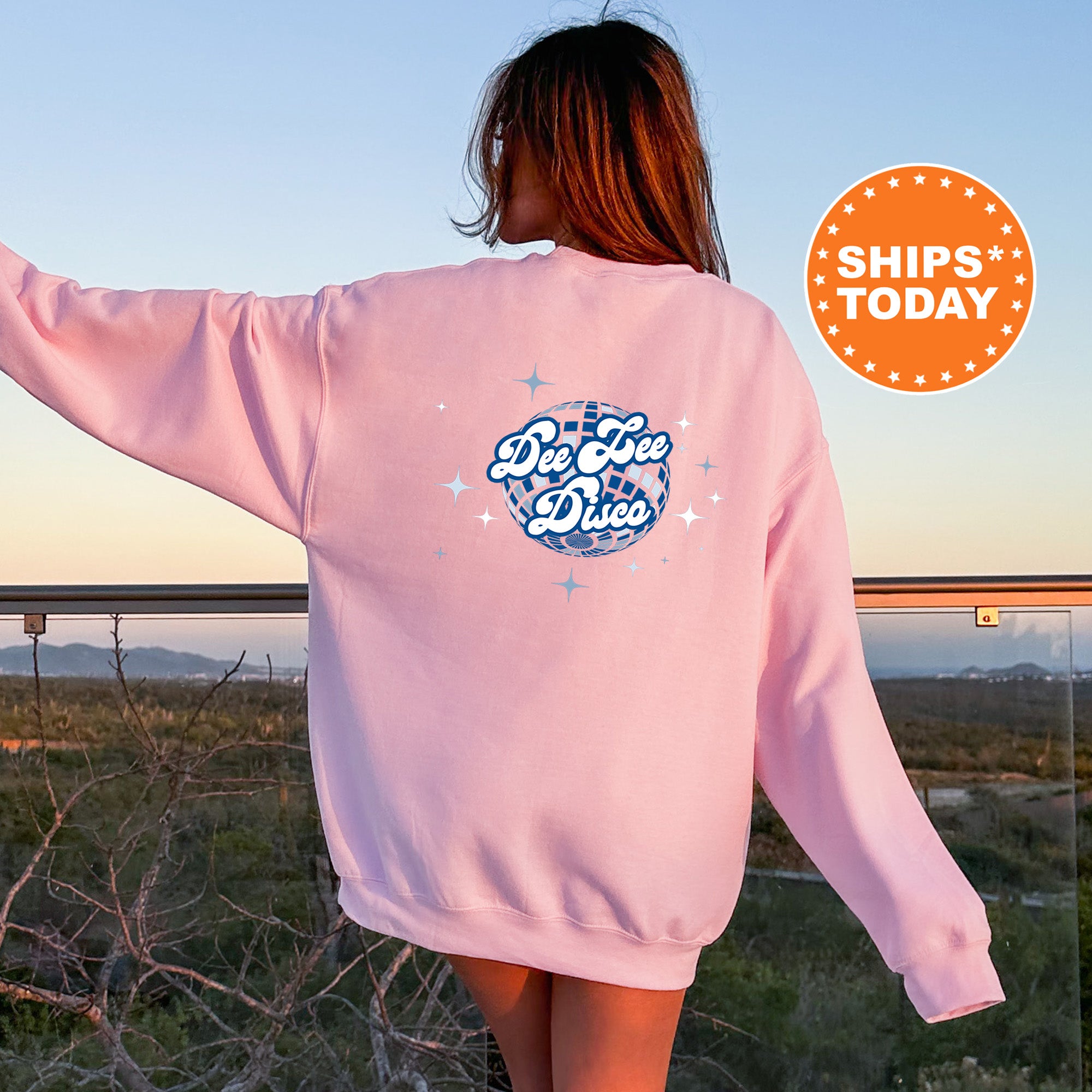 A woman wearing a pink sweatshirt with a graphic design on the back, standing in front of a scenic outdoor landscape with mountains in the distance.