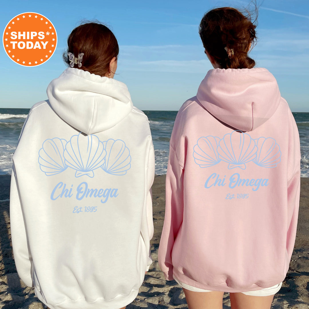 Two women wearing matching white and pink hoodies with the Chi Omega logo standing on a beach.