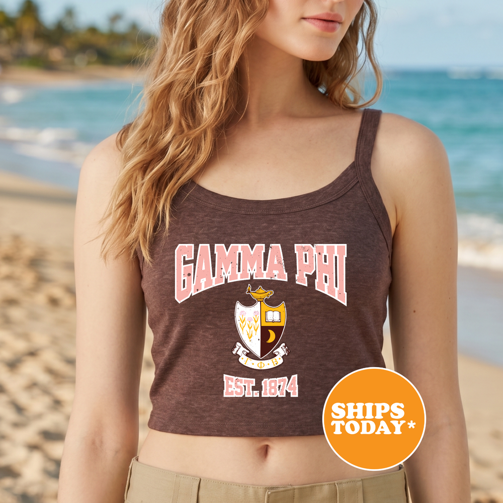 A woman wearing a brown tank top with the text "GAMMA PHI EST. 1874" printed on it, standing on a beach with palm trees in the background.