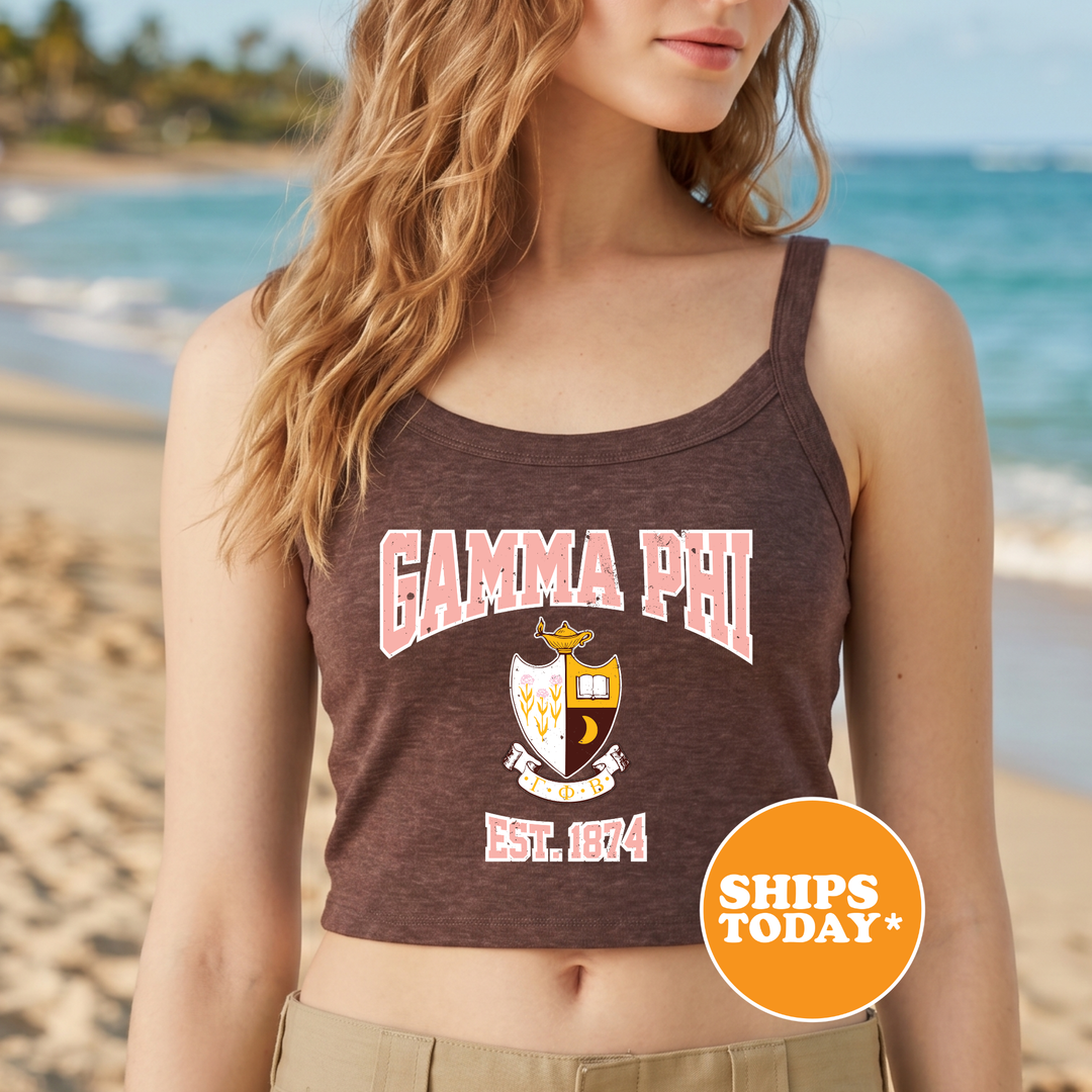 A woman wearing a brown tank top with the text "GAMMA PHI EST. 1874" printed on it, standing on a beach with palm trees in the background.