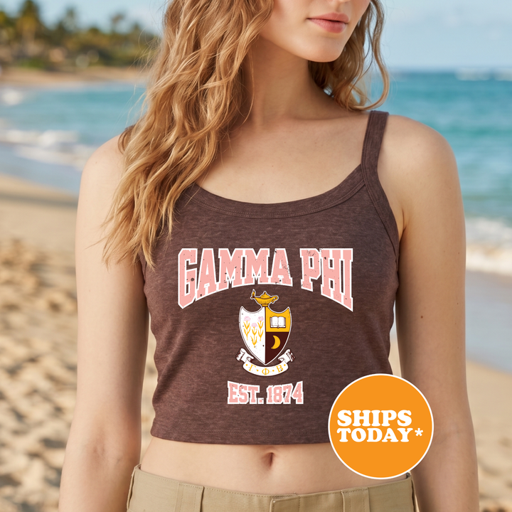 A woman wearing a brown tank top with the text "GAMMA PHI EST. 1874" printed on it, standing on a beach with palm trees in the background.