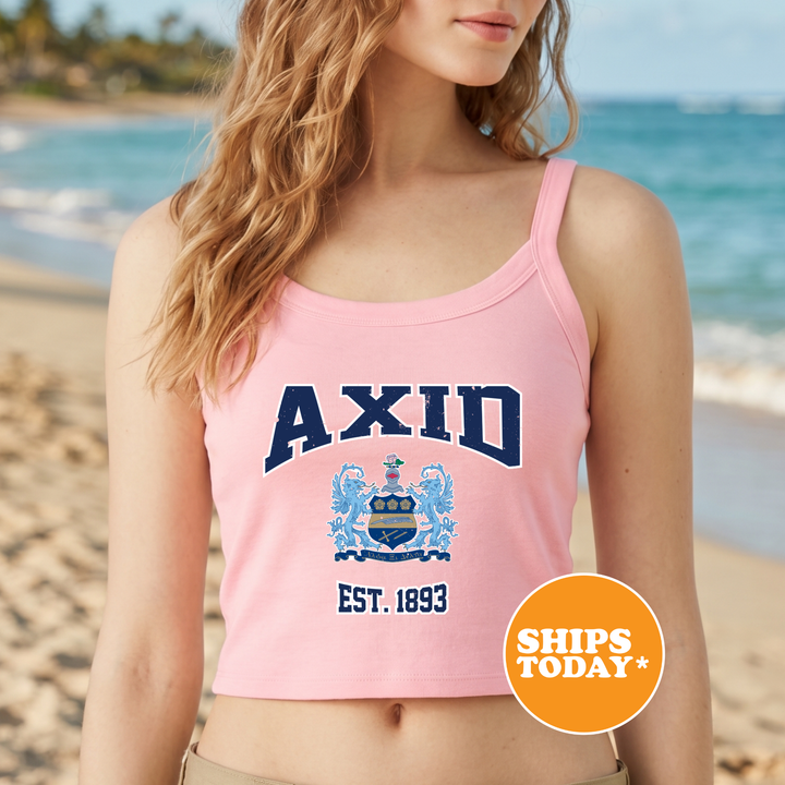 A woman wearing a pink crop top with the text "AXID EST. 1893" printed on it, standing on a beach with the ocean in the background.