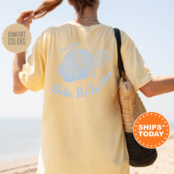 A person wearing a yellow Delta Phi Epsilon t-shirt with a seashell design, standing on a beach with a handbag.