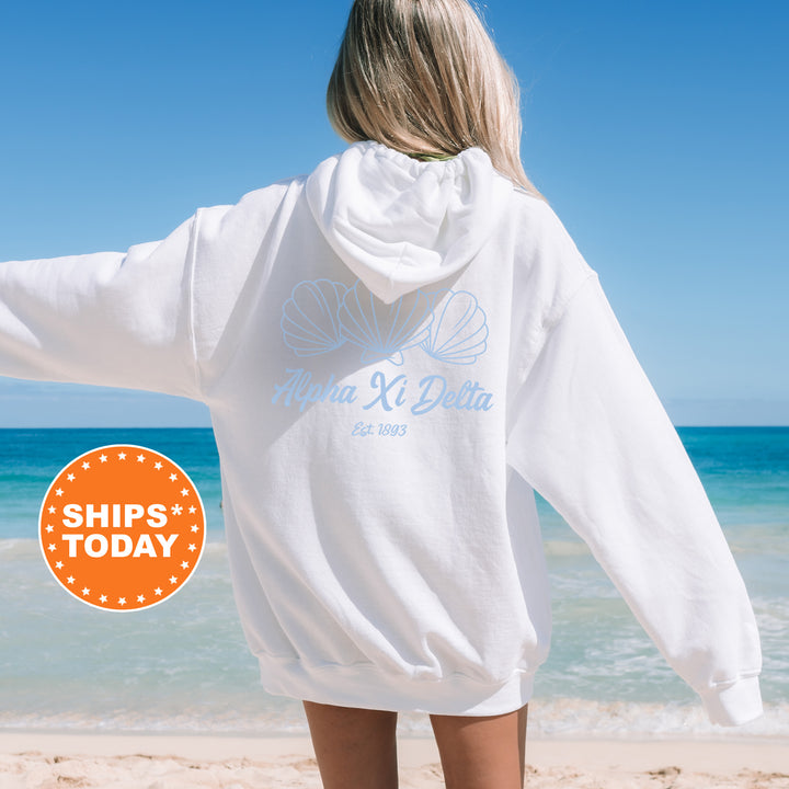 A woman wearing a white hoodie with the text "Alpha Kappa Delta" printed on it, standing on a beach with the ocean in the background.