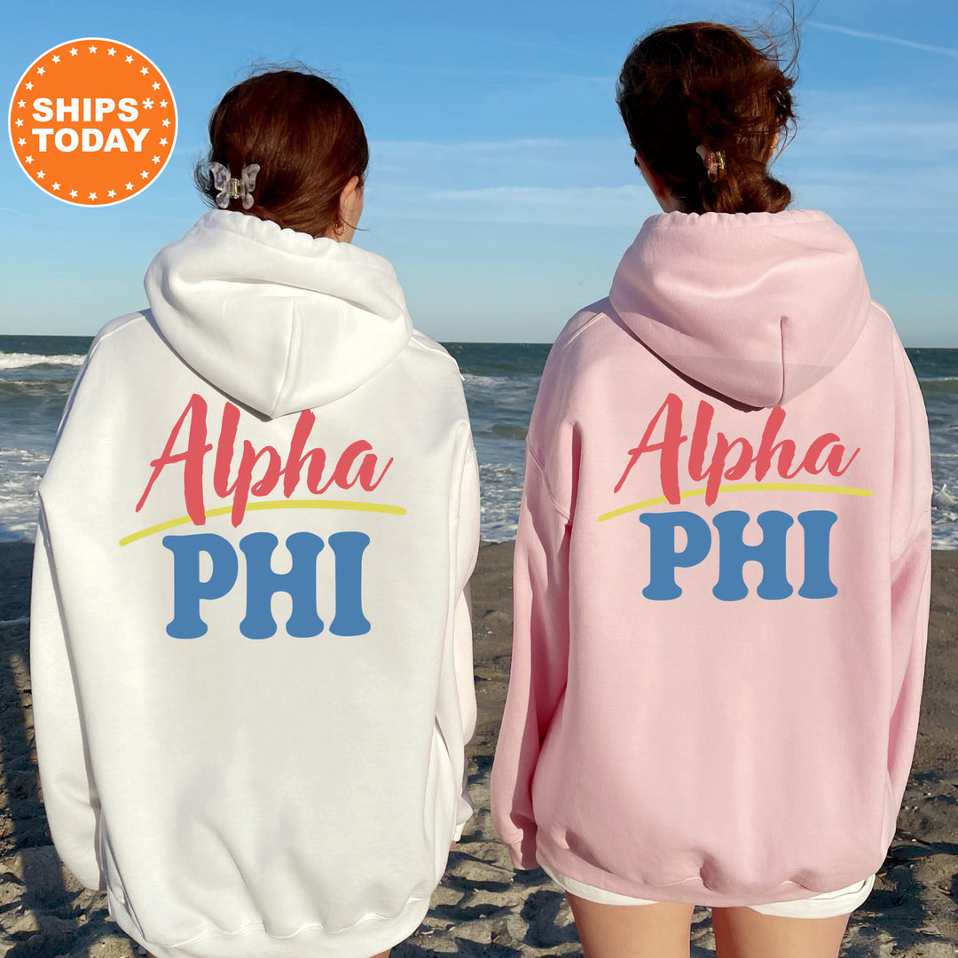 Two women wearing matching white and pink hoodies with the text "Alpha Phi" printed on them, standing on a beach with the ocean in the background.