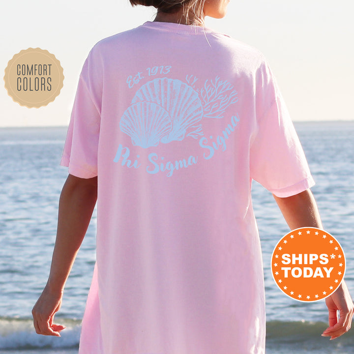 A person wearing a pink t-shirt with the text "Phi Sigma Delta" printed on it, standing on a beach with the ocean in the background.
