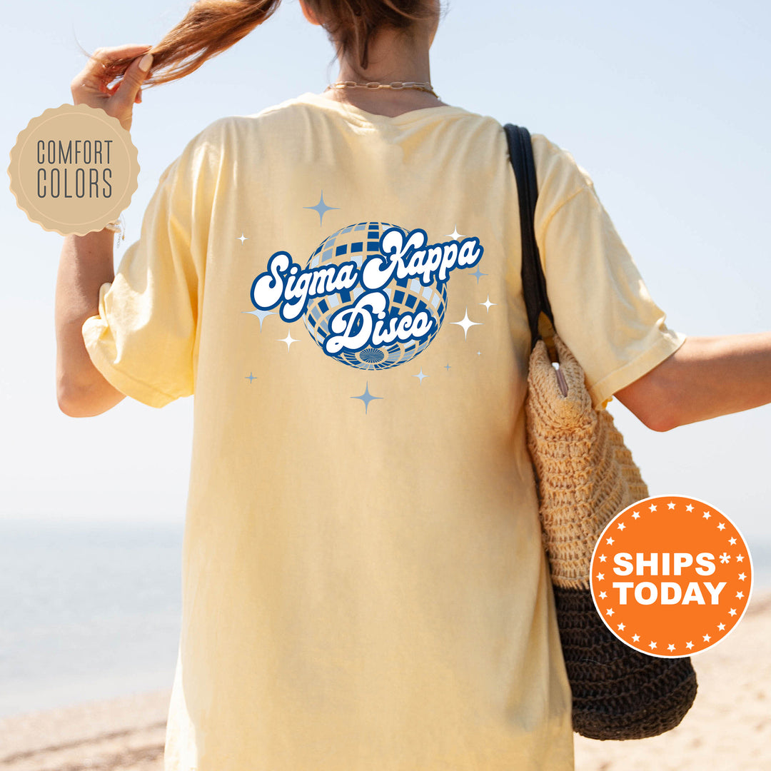 A person wearing a yellow t-shirt with the text " Sigma Kappa D disco" printed on it, standing on a beach with a handbag.